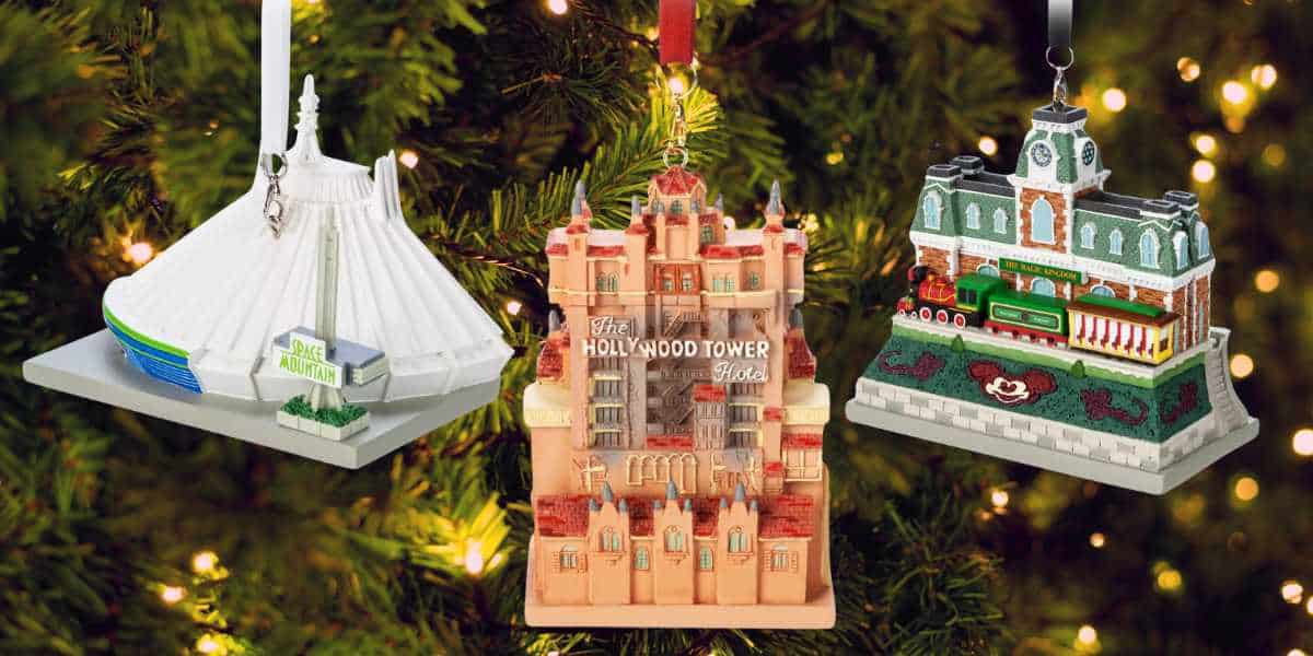 Three Disney Park-themed ornaments depict iconic attractions: a pyramid-shaped white building, a detailed multi-story hotel facade, and a colorful train station. They hang beautifully in front of a tree with twinkling lights.