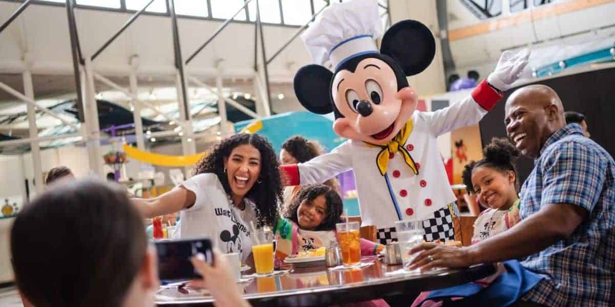 A family smiles and poses for a photo with a person dressed as a famous cartoon mouse in a chef outfit at Disney World. They are seated at a table with drinks and meals, enjoying the themed dining event in the brightly lit venue, complete with allergy information for added peace of mind.