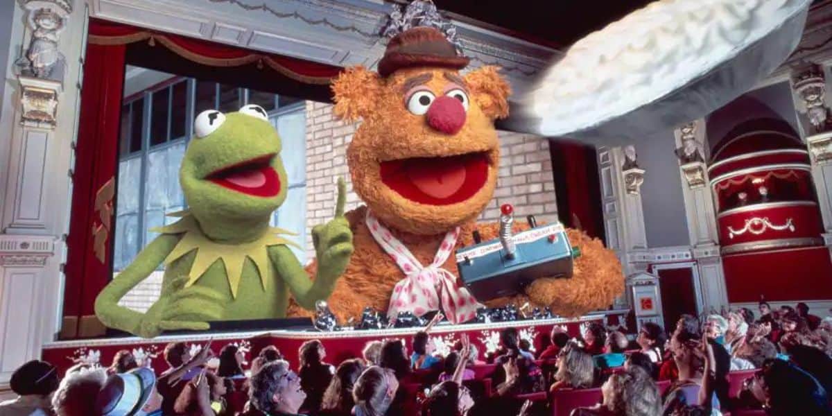 Kermit the Frog and Fozzie Bear from The Muppets delight families in a theater surprise. Amid sale rumors, Fozzie holds a pie while Kermit smiles. The scene is lively, with an ornate theater interior reminiscent of Jim Henson's legacy and enthusiastic viewers filling the seats.