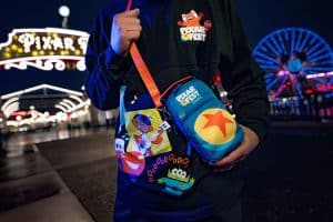 A person stands at an amusement park, wearing a Pixar Fest sweatshirt. They hold Pixar-themed bags from the Better Together Pixar Parade, featuring characters and designs from various films. Bright lights and a Ferris wheel glow joyfully in the background.