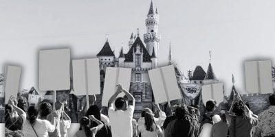 A group of people, seemingly on vacation, holds blank protest signs in front of a Disney castle. The fairytale structure's parapets and turrets contrast against the clear sky, blending black-and-white and colored elements to create an intriguing scene at Disneyland California as a Disneyland heightened security visit takes place.