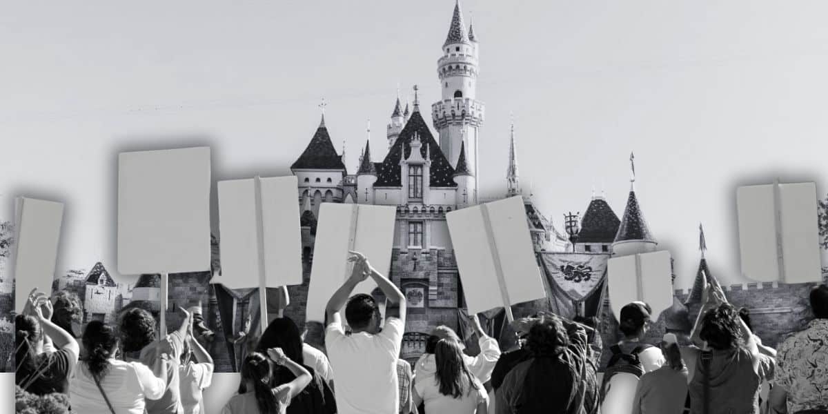 A group of people, seemingly on vacation, holds blank protest signs in front of a Disney castle. The fairytale structure's parapets and turrets contrast against the clear sky, blending black-and-white and colored elements to create an intriguing scene at Disneyland California as a Disneyland heightened security visit takes place.