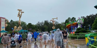People wearing rain ponchos walk through a theme park area with oversized toy decorations and a large Buzz Lightyear statue. The ground is wet, suggesting recent rainfall. Trees and park signs are visible in the background.