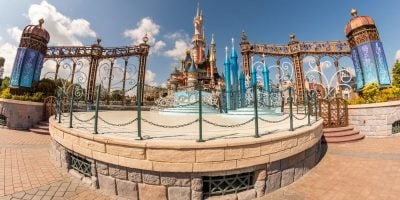 A fairy tale castle with ornate spires and turrets stands behind decorative gates, hosting eager theme park goers. The scene unfolds under a bright blue sky with scattered clouds, creating a whimsical and magical atmosphere reminiscent of Disney World but at this Disney Park, Disneyland Paris.