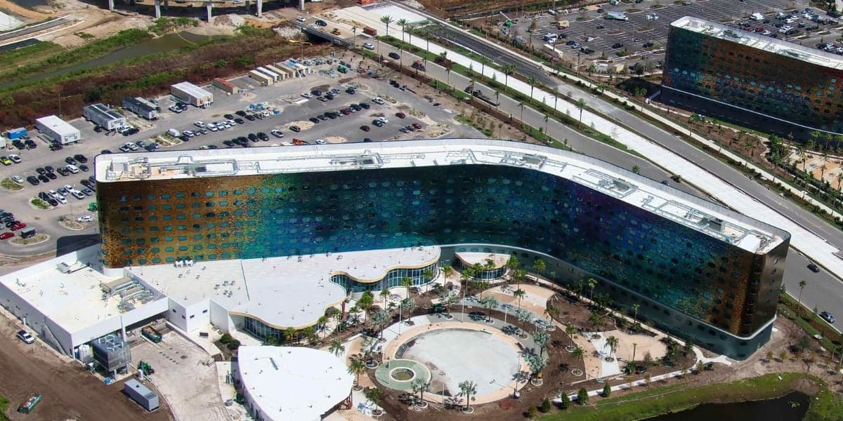 Aerial view of a large, modern building with a distinctive curved design, featuring colorful windows. Surrounding areas include parking lots, a circular landscaped area, and nearby roads.