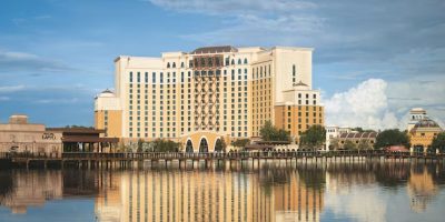 The tall hotel, with beige and brown accents, reflects beautifully on the calm lake. Its arches and numerous windows offer a picturesque view. Fluffy clouds decorate the blue sky while trees line the waterfront, making it hard to believe this is known as the worst hotel near Disney World.