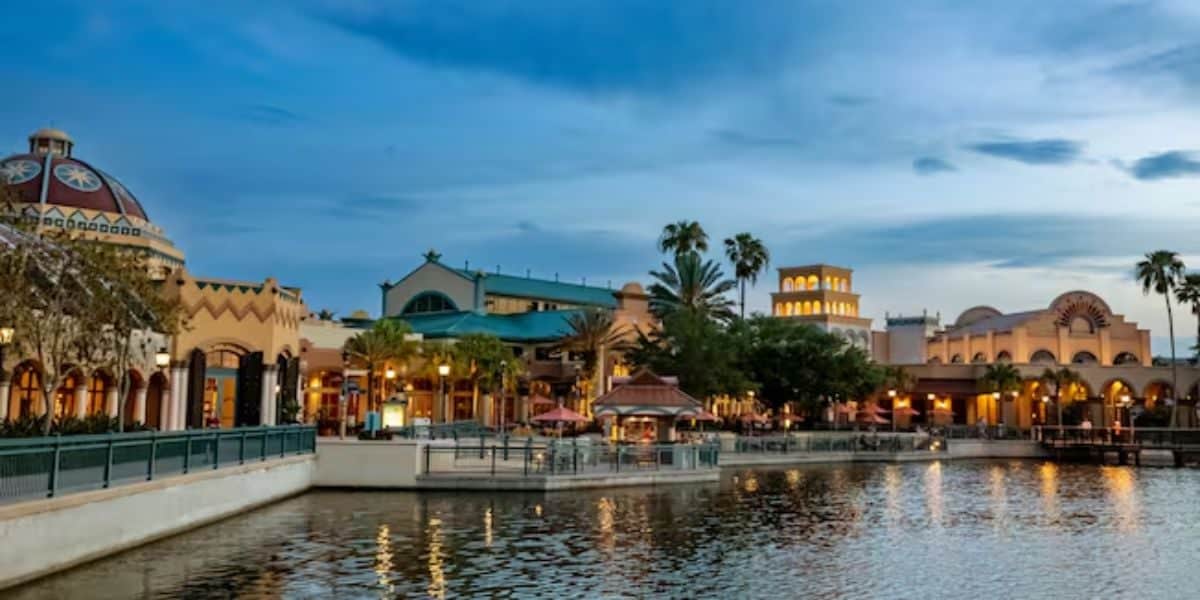 A picturesque waterfront with colorful, Mediterranean-style buildings illuminated at dusk evokes a sense of Disney World's magic. The scene reflects on the calm water, surrounded by palm trees under a blue, cloudy sky.