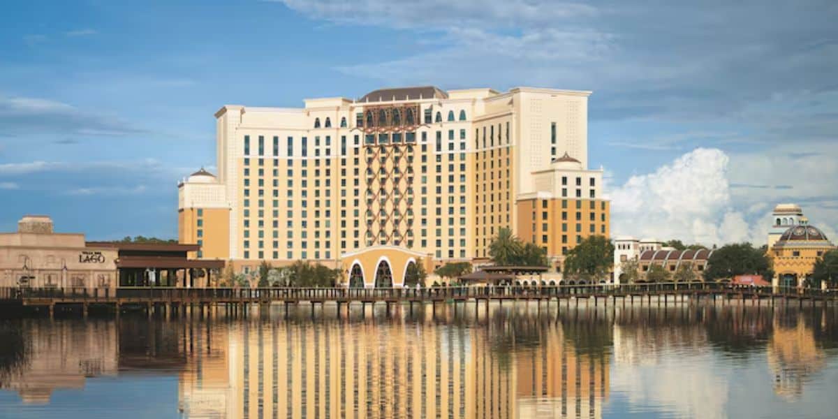 The tall hotel, with beige and brown accents, reflects beautifully on the calm lake. Its arches and numerous windows offer a picturesque view. Fluffy clouds decorate the blue sky while trees line the waterfront, making it hard to believe this is known as the worst hotel near Disney World.