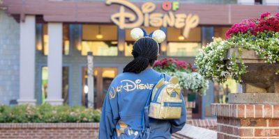 A person sporting Mickey Mouse ears and a blue Disney jacket stands facing a Disney store entrance, carrying one of those sought-after Disney Loungefly bags worth hundreds of dollars. Bright flowers adorn the foreground, adding to the vibrant scene.