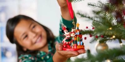 A young girl in a green patterned shirt decorates a holiday tree with a Disney Store ornament featuring a cartoon mouse in a festive outfit. The tree is adorned with red and gold ornaments, adding magic to the softly lit room. She smiles happily, captured by the festive charm.