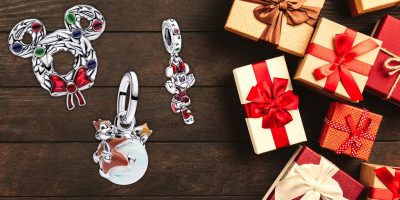 A festive scene with Disney-themed holiday ornaments, including a Mickey Mouse-shaped wreath and a Pandora character charm, arranged next to wrapped gifts with red ribbon bows on a wooden surface from the Disney Store.