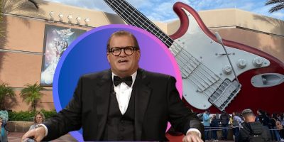 A man in a tuxedo and glasses stands in front of an enormous guitar sculpture at a Disney World attraction, with onlookers nearby. The backdrop features palm trees and a building with a musical theme, capturing the whimsical spirit of Disney.