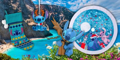 A scenic beach backdrop features festive holiday-themed Stitch items: a colorful Christmas stocking, a Disney Stitch ornament, a figurine holding a tree, and a circular backdrop with Stitch and friends, adding whimsical charm to the coastal view.