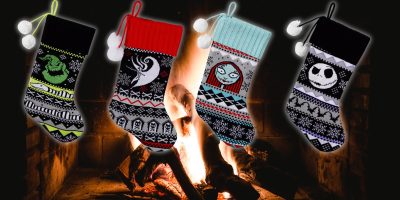 Four "Nightmare Before Christmas" stockings with spooky-themed designs hang in front of a cozy fireplace. Each stocking features different black and white patterns and stylized faces or motifs, set against the glow of a festive orange fire.