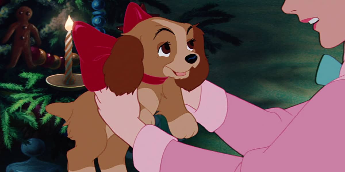 An animated scene shows a person holding a small brown puppy with long ears and big eyes. The puppy, part of the Disney Dogs Holiday Collection, has a red collar and large red bow. It stands in front of a decorated Christmas tree, with a lit candle and gingerbread man ornament visible in the background.