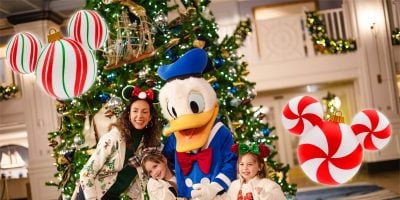 A woman and two children pose with a character in a blue hat in front of a decorated Christmas tree. Large, festive ornaments shaped like Mickey Mouse ears are overlaid in the image, capturing the essence of Disney Magic.
