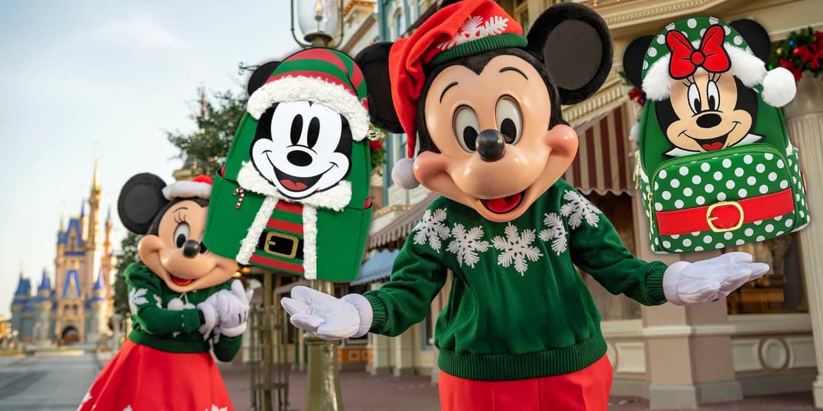 Mickey and Minnie Mouse, donning festive Christmas sweaters and hats, stand in front of a decorated castle. They hold Loungefly Holiday Elf Mini Backpacks adorned with their faces, smiling and waving joyfully.