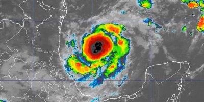A weather satellite image captures Hurricane Milton swirling over a landmass. The storm's eye is clearly visible at the center, encircled by vibrant red, orange, yellow, and green cloud patterns signaling intensity. Coastlines and geographical outlines are discernible in the background.