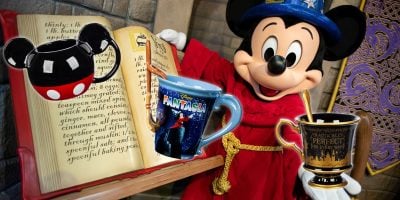 Mickey Mouse, dressed as a sorcerer, stands next to a large open book with a decorative background. In front of the book, three themed coffee mugs beckon every fan, capturing the magic one can only find at the Disney Store.