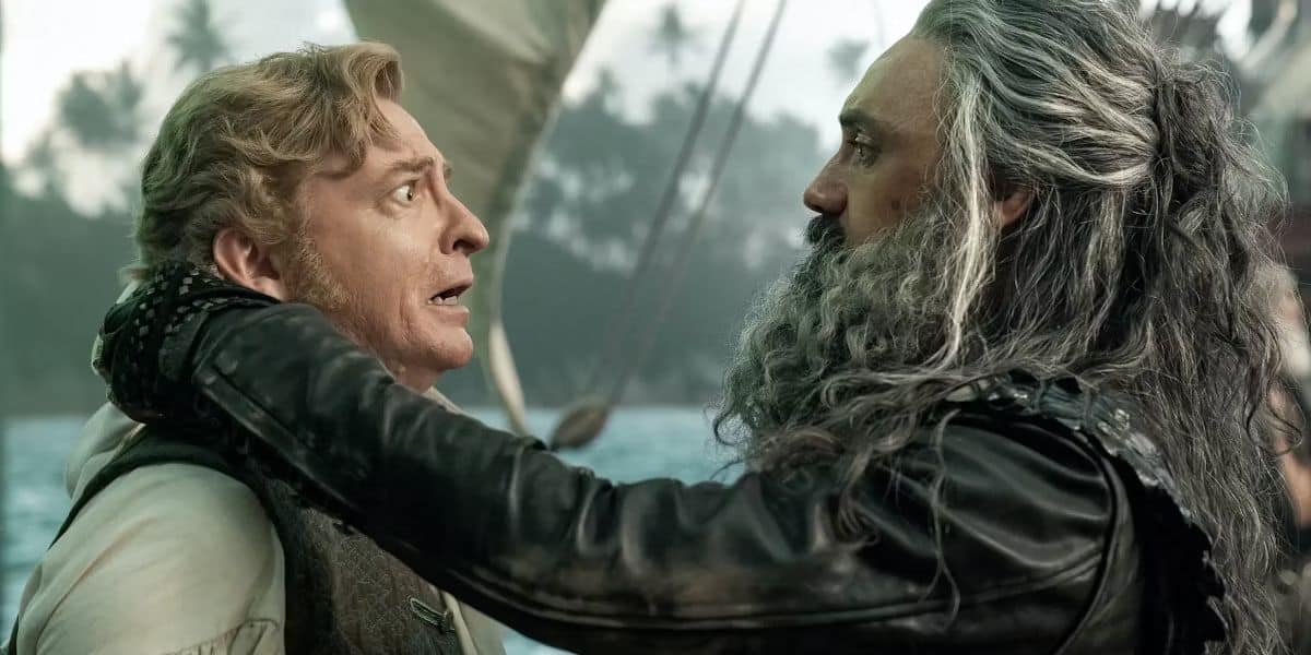 A scene with (left to right) Rhys Darby as Stede Bonnet and Taika Waititi as Edward Teach/Blackbeard; Bonnet with a concerned expression and light hair, Teach resembling a seasoned pirate with long gray hair and beard, gently holding Bonnet's shoulders. The background shows sails billowing against a misty shoreline dotted with trees.