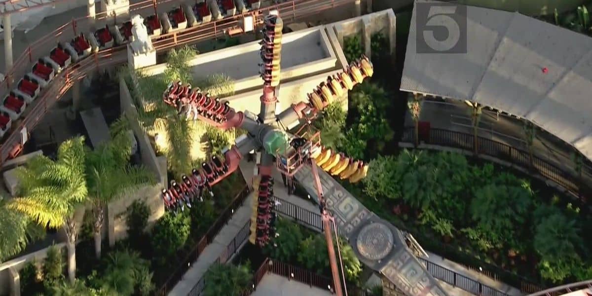 Aerial view of a rotating amusement park ride with multiple arms, surrounded by lush greenery and pathways. The ride seats are filled with people, and there's a nearby section of a roller coaster track, inside a California theme park.