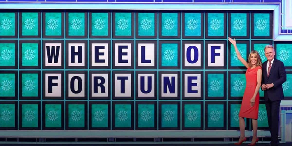 A woman in a red dress points to a game board displaying "WHEEL OF FORTUNE" in blue and white tiles. A man in a suit stands beside her, smiling. The background is a vibrant blue with decorative patterns.