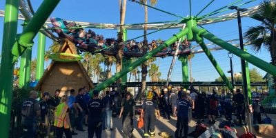 A green roller coaster with passengers is stalled mid-ride. Emergency responders and park staff, wearing uniforms and safety gear, gather below to assist. Palm trees and a clear blue sky are in the background.