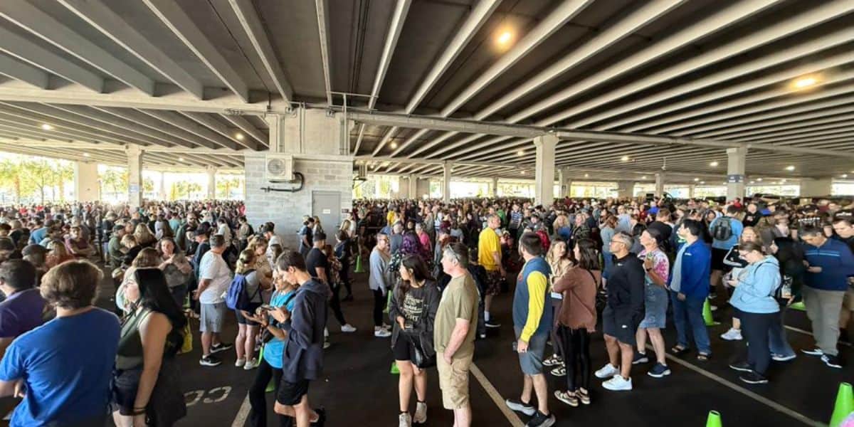 A large crowd of people gathered under a concrete overpass, standing in multiple lines. Many individuals are casually dressed and the area is filled with a mix of ages and genders. Some people are using their phones while waiting.