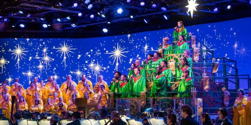 A vibrant choir sings on a tiered stage, dressed in green and yellow robes under star-like lights. A conductor leads the orchestra in the foreground, creating a festive and joyful atmosphere.