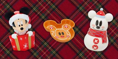 Festive holiday scene brimming with Disney magic: a Mickey Mouse figurine in a Santa hat bursts from a gift box, accompanied by a Mickey-shaped gingerbread dish and a snowman figurine sporting Mickey ears, scarf, and hat—all set against a vibrant red plaid background.