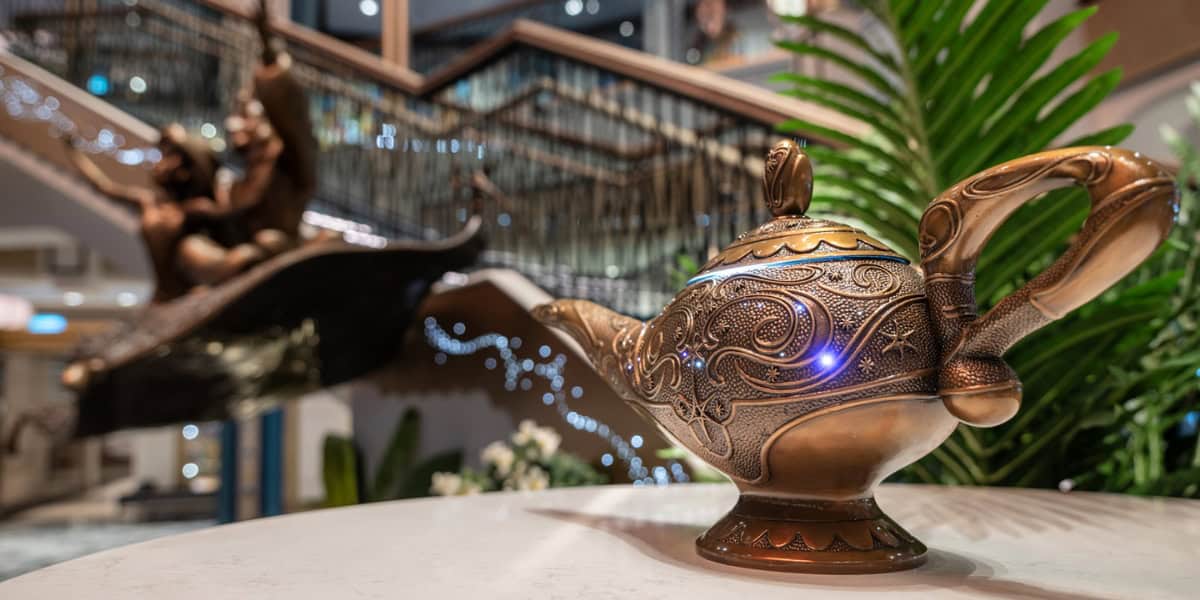 A decorative bronze teapot with intricate engravings sits prominently on a marble table, evoking the enchanting charm of Disney Treasure. In the background, a blurred statue portrays a person on a carpet, suggesting magic. Green plants and a staircase add depth to this captivating scene.