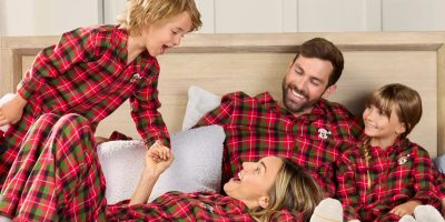 A family of four, clad in matching red plaid Disney pajamas, is sitting on a bed.