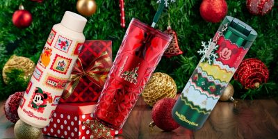 Three festive Starbucks holiday drinkware containers are displayed in front of a decorated Christmas tree with ornaments and gifts. The containers feature colorful patterns and beloved seasonal motifs, perfect for spreading holiday cheer.