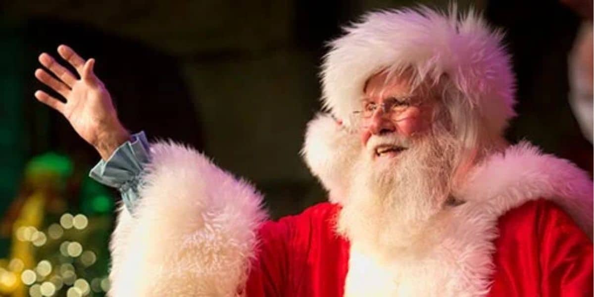 A person in a Santa Claus costume with a fluffy white beard and red suit waves cheerfully. The background is softly blurred, with warm, festive lighting creating a joyful ambiance, inviting guests to meet Santa amidst the holiday magic.