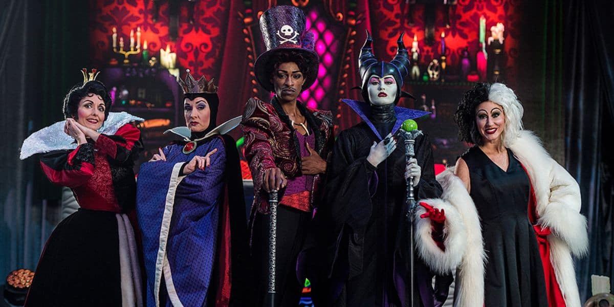 A group of five people dressed as various fairy tale villains stands in a theatrical setting reminiscent of Villains Land, with a dark, ornate background. They wear elaborate costumes and makeup, each with unique and distinctive details that capture the magic from Disney's enchanted realms.