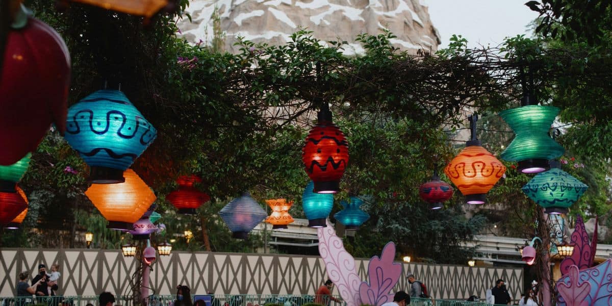 Colorful lanterns hang from a vine-covered structure, casting a magical Disney ambiance in an outdoor setting. A mountainous structure looms in the background, and people, perhaps gathered from Disney fan clubs, enjoy the lively atmosphere below.