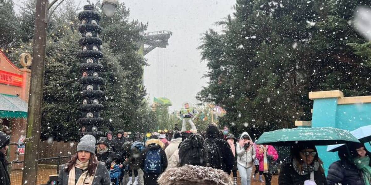 A crowded outdoor area at a Disney Park during snowfall creates a magical Winter Sight. People are bundled up in winter clothing, some holding umbrellas as snowflakes dance in the air. In the background, trees and a towering structure are partially covered in snow.