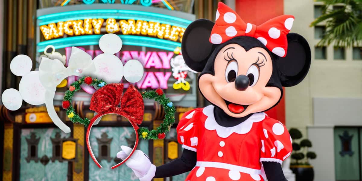 A character in a red polka-dot dress and bow, reminiscent of Minnie Mouse, stands outside a colorful theater entrance. A pair of light-up ears is prominently displayed in the festive foreground.