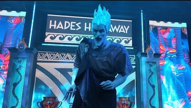 At Oogie Boogie Bash 2025, a performer dressed as Hades from Disney's Hercules stands on stage with blue flaming hair and dark robes. The backdrop features Greek-style motifs and the words "Hades Hangout," while blue and orange lighting create an intense atmosphere.