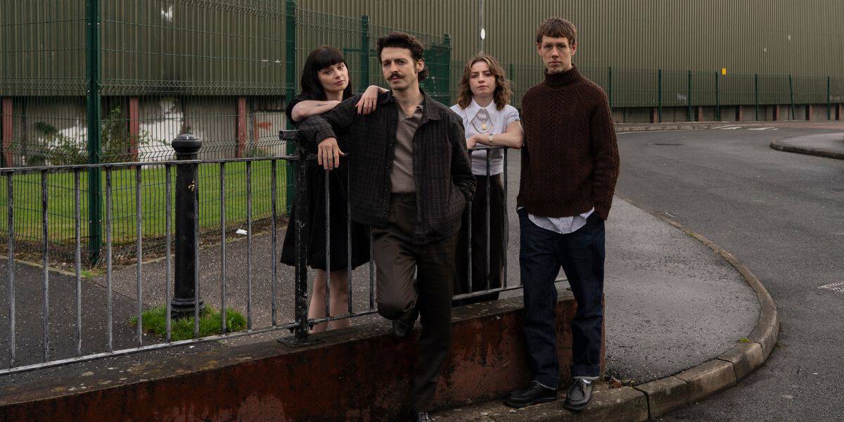 Four people stand outdoors near a metal fence by a street. Two lean on the fence, while two stand close by. They are casually dressed, and the background includes a large industrial building and a paved road. The scene is overcast.
