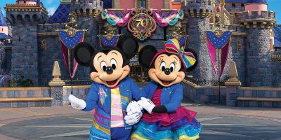Mickey and Minnie Mouse, wearing colorful outfits, stand hand-in-hand in front of a castle decorated for a 70th anniversary celebration of this Disney Park.
