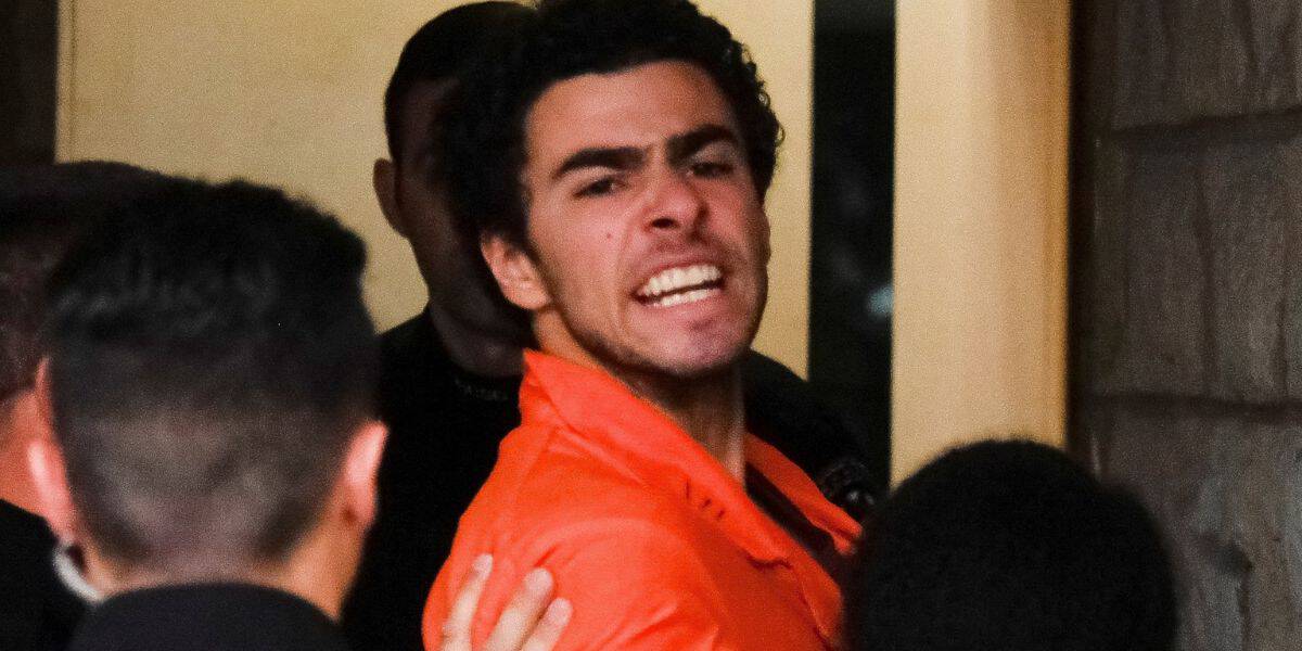 A man in an orange shirt appears distressed and is being held by several individuals, possibly security or law enforcement, in a dimly lit environment.