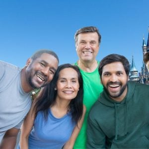 A group of Disney Adults smiling and taking a selfie in front of the Cinderella Castle in Magic Kingdom. The sky is clear and blue in the background, suggesting a sunny day at Disney World.