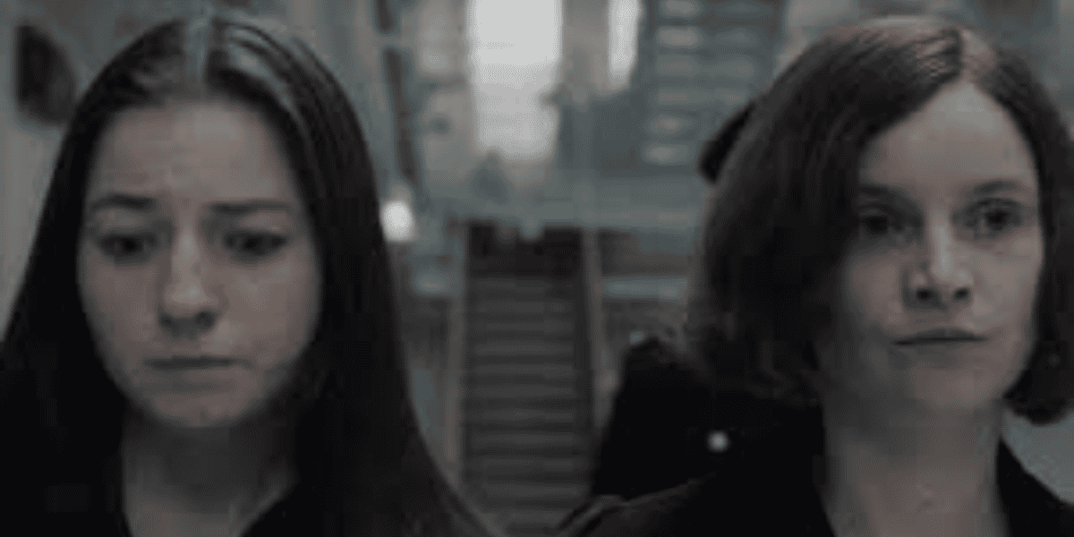 Two women walking indoors with serious expressions. The woman on the left has long dark hair, and the woman on the right has short, reddish-brown hair. The background shows a blurred staircase and a figure in uniform.