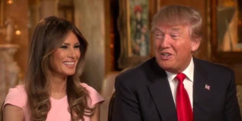 A smiling woman with long brown hair wearing a pink dress sits next to a man with blond hair, reminiscent of a Disney prince, wearing a suit, white shirt, and red tie. They are indoors, in an elegantly decorated setting.