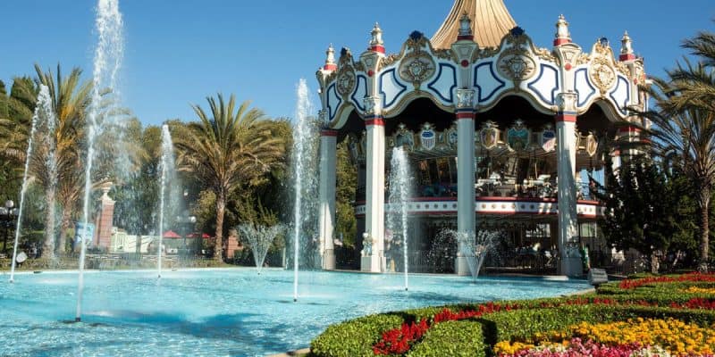 A large, ornate carousel near a sparkling fountain, surrounded by palm trees and vibrant flower beds under a clear blue sky.