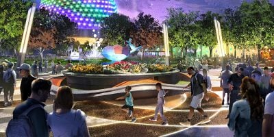 People stroll through a vibrant, futuristic park at night, illuminated by colorful EPCOT World Celebration lights. A glowing geodesic dome stands in the background as beams of light dance in the sky. With trees and a water fountain, the lively atmosphere enchants all who visit at Disney World.