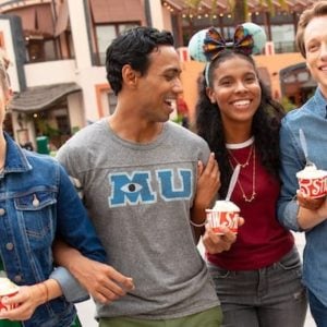Four friends, looking like Disney cast members on their day off, stroll outdoors with smiles and cups of frozen yogurt. Casually dressed, they revel in the sunny day at an amusement park or bustling outdoor shopping area, with buildings and trees framing their joyful scene.