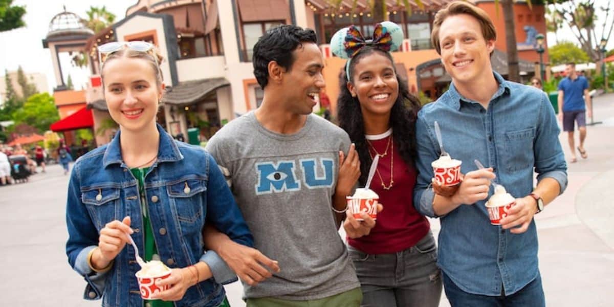 Four friends, looking like Disney cast members on their day off, stroll outdoors with smiles and cups of frozen yogurt. Casually dressed, they revel in the sunny day at an amusement park or bustling outdoor shopping area, with buildings and trees framing their joyful scene.