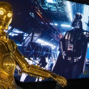A gold humanoid robot stands in the foreground, while a large screen in the background displays a black-armored figure with a cape, surrounded by troops in white armor inside a space hangar, reminiscent of an epic scene you might encounter on a Disney World Vacation.